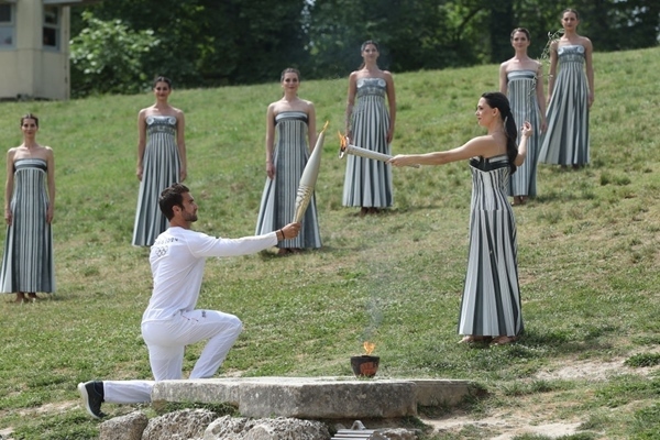 扮演最高女祭司的演員瑪麗·米娜（前右）點(diǎn)燃第一棒火炬手——希臘運(yùn)動(dòng)員、東京奧運(yùn)會(huì)男子賽艇項(xiàng)目金牌得主斯特凡諾斯·恩圖斯科斯手中的火炬。新華社記者 趙丁喆 攝